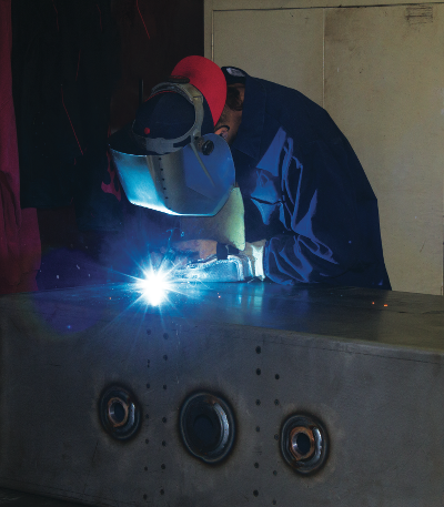 The image shows a person bent over a steel reservoir while welding together the sides. A light blue spark can be seen in the photo that is also reflecting off of the reservoir and the welding helmet the person is wearing.