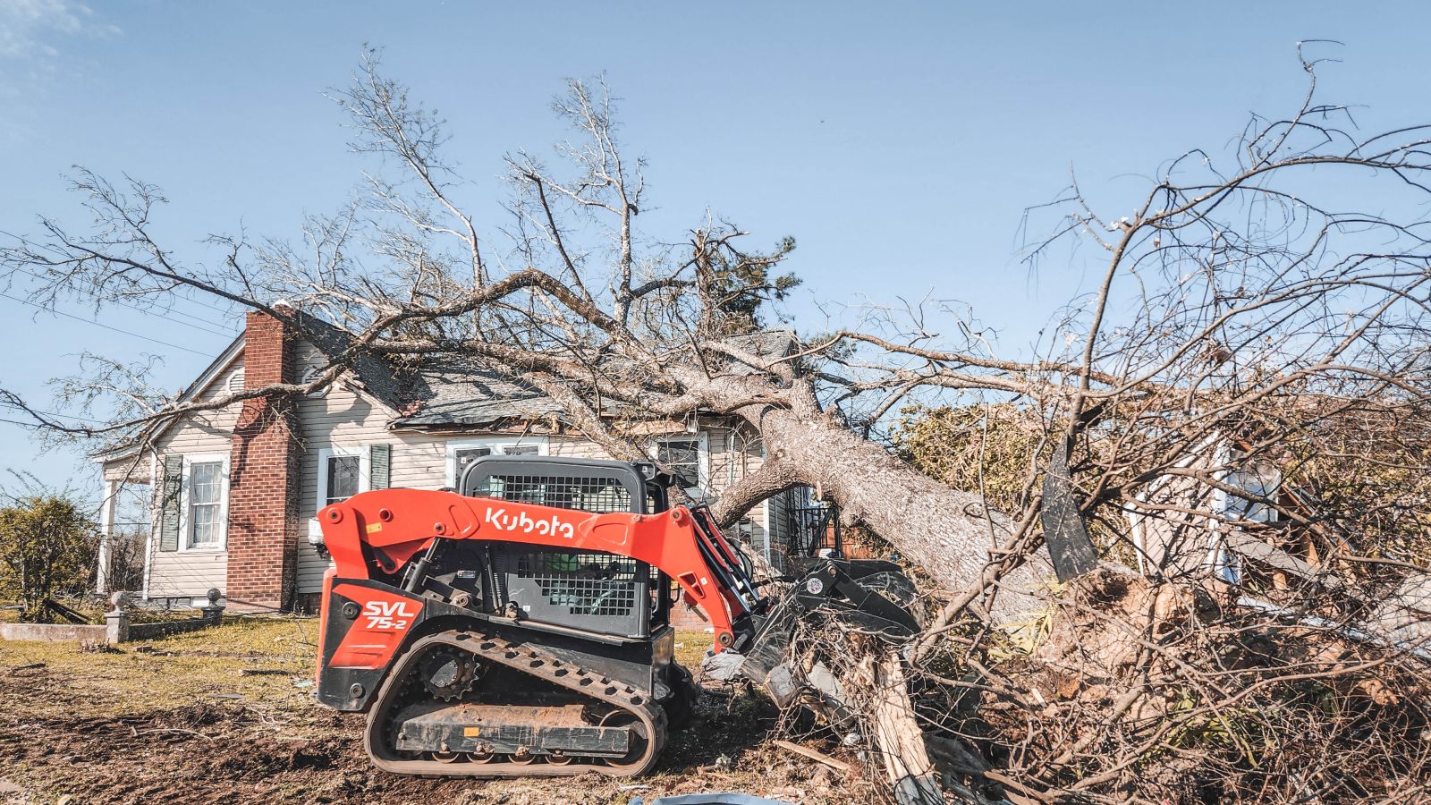 The image shows a tree fallen on a house and a skid steer near the base of the tree.